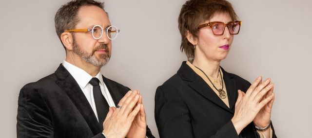 Greg Wilder and Alison Wilder are standing together, hands together in either prayer mode or evil genius mode. Hard to tell.