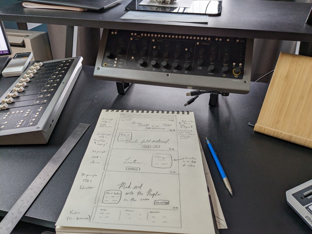 Messy black desk with audio equipment cleared out of the way to make room for a sketchpad with pencil-drawn website wireframes 
