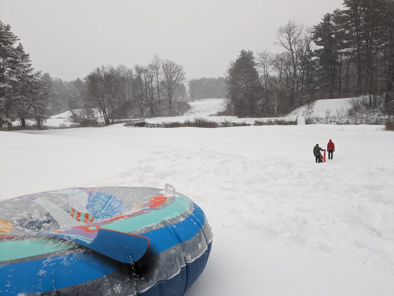 A snowy field with a inner tube in the foreground and adults and a child down the hill in the distance